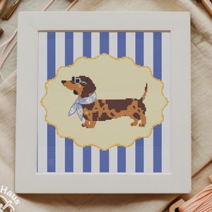 May include: A framed cross-stitch featuring a dachshund wearing goggles and a scarf, set against a blue and white striped background. The dog is brown and tan with a yellow border. The frame is white.