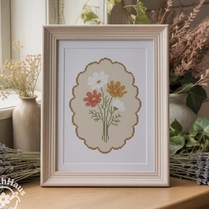 May include: A framed cross-stitch embroidery featuring a bouquet of white, pink, and orange cosmos flowers centred within a scalloped oval border on a beige background. The artwork is encased in a light-coloured wooden frame with vertical grooves.
