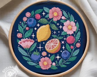 May include: A cross-stitch embroidery hoop featuring a botanical design. The design includes a yellow lemon, a sliced grapefruit, pink flowers, blueberries, and green leaves on a dark blue background. The wooden hoop is approximately 8 inches in diameter.