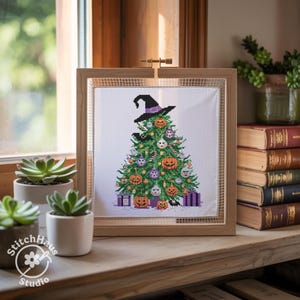 May include: A framed cross-stitch design featuring a Halloween-themed Christmas tree. The tree is decorated with jack-o'-lanterns, purple ornaments, and a witch's hat. The frame is made of light-colored wood. The StitchHaus Studio logo is visible.