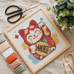 Peut inclure: Point de croix d'un chat porte-bonheur rouge et jaune tenant une pièce d'or, encadré en bois clair. Comprend des ciseaux noirs, du fil à broder de différentes couleurs et une petite plante.