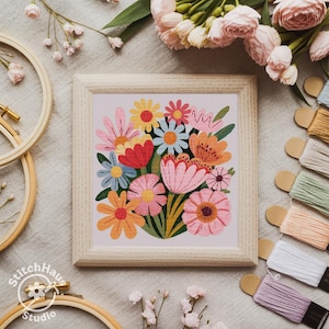 May include: A framed cross-stitch floral design with a bright bouquet of flowers in pink, orange, yellow, red, and blue. The frame is light wood, and the design is surrounded by embroidery threads and floral accents.