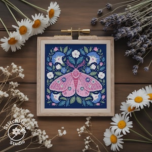 Pode incluir: Uma peça de ponto de cruz emoldurada com uma mariposa rosa com padrões intrincados nas asas, rodeada por elementos florais azuis e brancos sobre um fundo azul marinho. A obra está num bastidor de madeira, com o logótipo StitchHaus Studio visível.