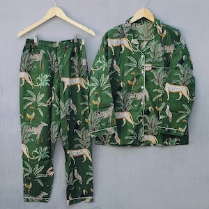 Puede incluir: Conjunto de pijama verde con estampado de animales de la selva, incluyendo cebras y leopardos. La parte superior de manga larga y los pantalones son de tela suave y tienen ribetes blancos. El conjunto está colgado en una percha de madera.