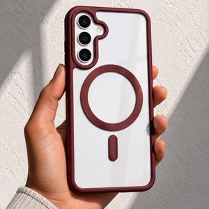 May include: A clear smartphone case with a deep burgundy frame and a matching circular magnetic charging ring on the back. The case features a cutout for a triple-lens camera system and a small vertical alignment bar below the magnetic ring.