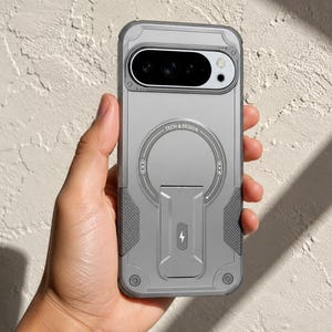 May include: A matte grey smartphone case featuring a prominent circular magnetic ring with the text TECH & DESIGN printed along the top. The case includes a foldable kickstand in the centre with a lightning bolt icon and textured side grips.