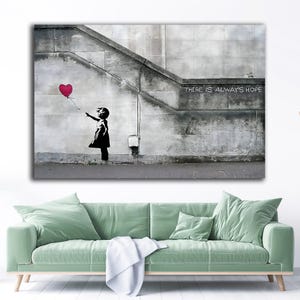 May include: A canvas print of a Banksy artwork. A black silhouette of a child reaching for a red heart-shaped balloon. The background is a grey concrete wall with the text "THERE IS ALWAYS HOPE".