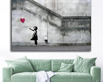 May include: A canvas print of a Banksy artwork. A black silhouette of a child reaching for a red heart-shaped balloon. The background is a gray concrete wall with the text "THERE IS ALWAYS HOPE".