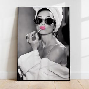 Può includere: Stampa incorniciata in bianco e nero di una donna che applica il rossetto. Indossa occhiali da sole, un asciugamano bianco e un accappatoio bianco. Il rossetto è rosa acceso, creando un contrasto con l'immagine in scala di grigi.