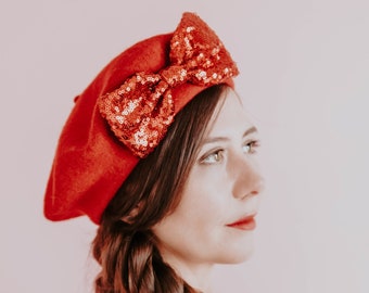 Könnte beinhalten: Ein rotes Barett mit einer großen Pailletten-Schleife. Das Barett hat eine kräftige, rote Farbe, und die Schleife besteht aus roten Pailletten. Der Hut ist ein stilvolles Accessoire.