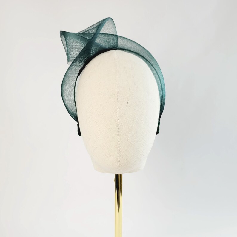 May include: A dark green tulle headband with a twisted design. The headband is on a white mannequin head.
