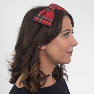 May include: A red plaid bow headband. The bow features a classic tartan pattern with green, yellow, and white accents. The headband is worn on a person with dark brown hair and a black top.