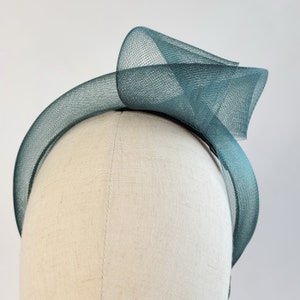 May include: A teal green mesh headband with a large bow on top. The headband is on a white mannequin head.