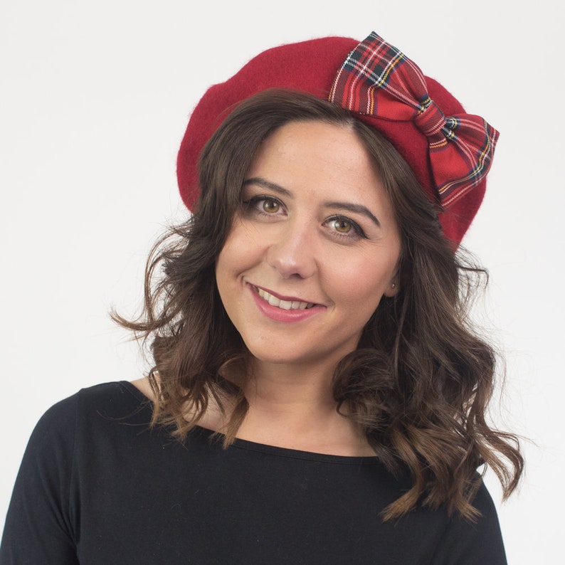 May include: A red beret with a plaid bow. The beret is a solid, deep red color, and the bow features a classic red, green, and yellow plaid pattern. The beret is worn by a person with brown hair and a black top.
