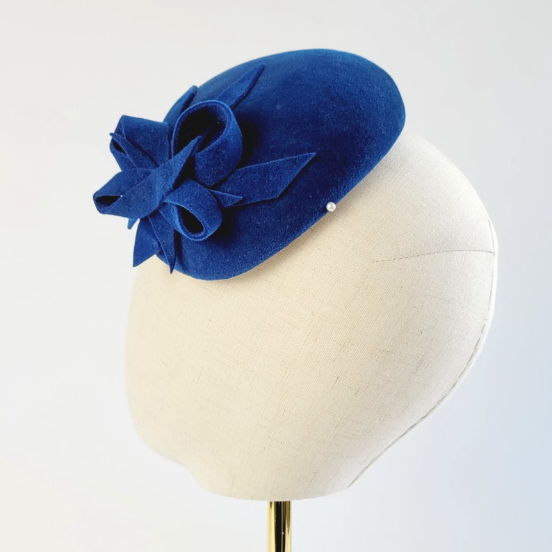 May include: A blue felt hat with a large blue felt bow on top. The hat is sitting on a white hat form.