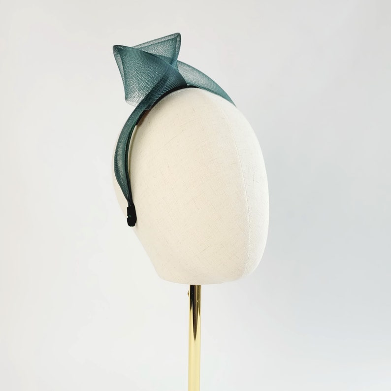 May include: A dark green fabric headband with a twisted design. The headband is on a white mannequin head.