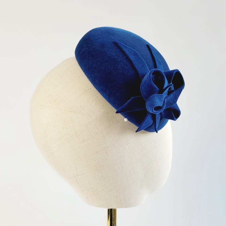 May include: A blue felt beret with a large blue felt flower accent. The beret is on a white mannequin head.