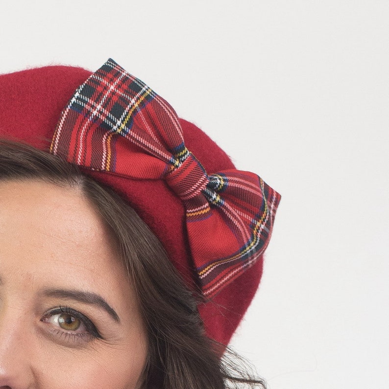 May include: A red beret with a plaid bow. The beret is a solid, deep red color, and the bow features a classic red, green, yellow, and white plaid pattern. The beret is a stylish accessory.
