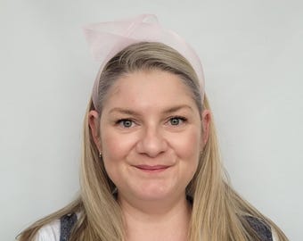 May include: A woman with long blonde hair is wearing a pink headband with a bow on top. The headband is made of a soft, delicate material.