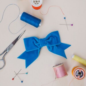 May include: A blue velvet bow with a pair of scissors, spools of thread, and sewing pins on a light beige background.
