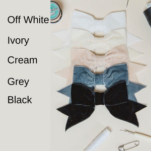 May include: Five velvet bows in different colors: off-white, ivory, cream, grey, and black. The bows are arranged in a stack, with the off-white bow on top and the black bow on the bottom.