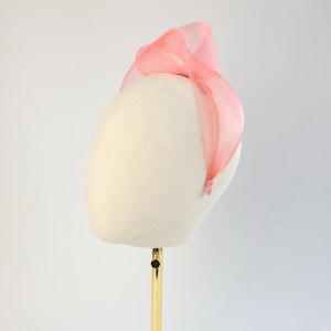 May include: A coral-coloured headband with a mesh texture, displayed on a mannequin head. The headband has a looped design at the top and a thin band that curves around the head. The background is plain white.