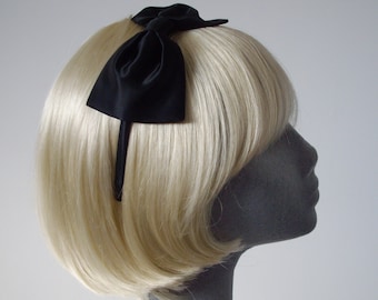 May include: A black satin bow headband. The headband is worn on a mannequin with blonde hair.