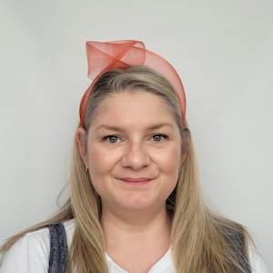 May include: A woman wearing a coral-coloured headband with a decorative bow. The headband is made of a sheer, textured material and sits atop the woman's blonde hair. She is wearing a white shirt and denim overalls.