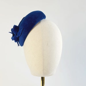 May include: A blue felt hat with a flower accent. The hat is on a white hat form.