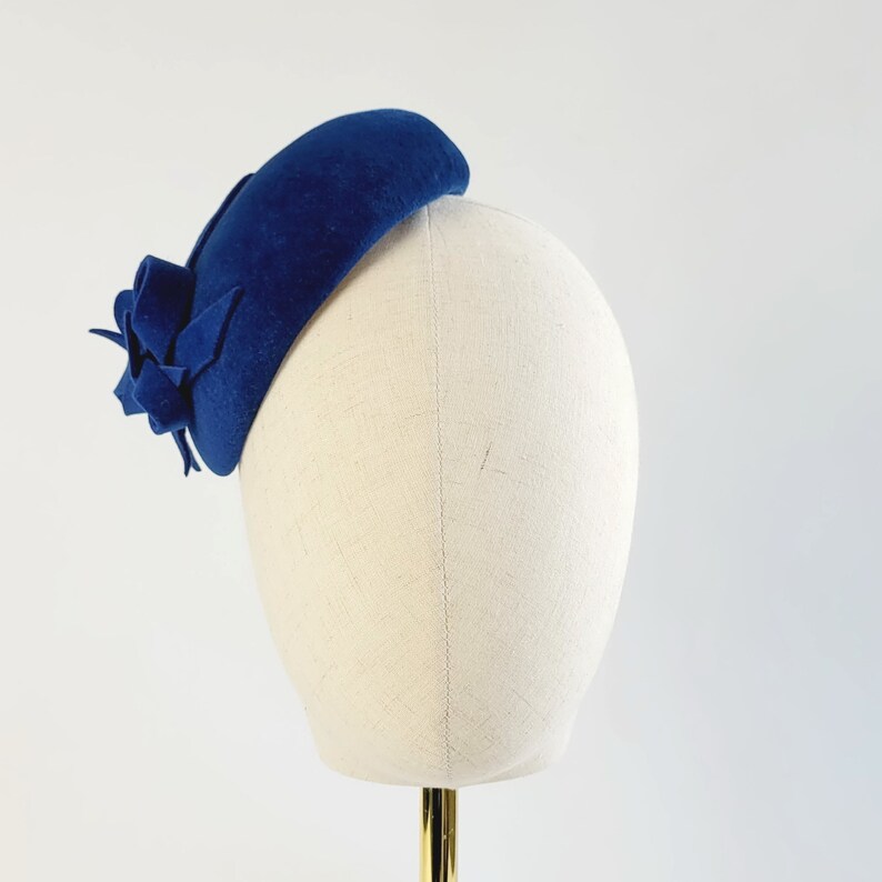 May include: A blue felt hat with a flower accent. The hat is on a white hat form.