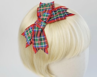 May include: Red and green plaid bow headband. The bow is tied in a large knot and has a pointed shape.