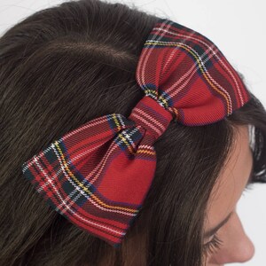 May include: A red plaid bow headband. The bow features a classic tartan pattern with red, green, yellow, and white stripes. The headband is worn on dark brown hair.