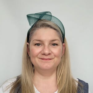 May include: A woman with long blonde hair is wearing a dark green headband with a bow on top. The headband is made of a soft, delicate fabric.