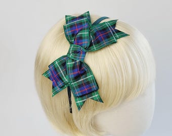 May include: A green, blue, and black plaid bow hair accessory. The bow is attached to a thin, dark headband. The bow is made of fabric and is styled on a mannequin head with blonde hair.
