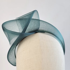 May include: A dark green,  mesh headband with a large,  twisted bow on top. The headband is on a white mannequin head.