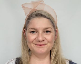 May include: A woman wearing a peach-colored headband with a large bow on top. The headband is made of a soft, sheer fabric.