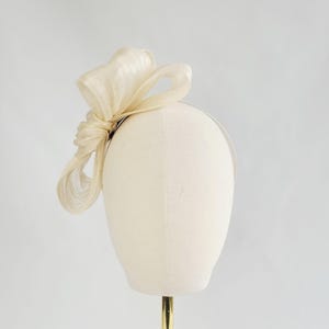 May include: A cream-colored fascinator with a large, knotted bow. The bow is made of a sheer, textured fabric and is attached to a thin, silver headband. The headpiece is displayed on a mannequin head against a white background.