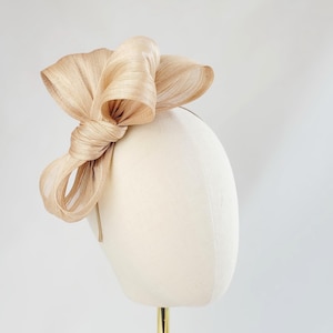 Peut inclure: Coiffe beige avec un grand nœud noué. Le nœud est fait d'un matériau tissé texturé et est fixé à un fin bandeau. La coiffe est présentée sur une tête de mannequin. Cet accessoire élégant est adapté aux occasions spéciales.