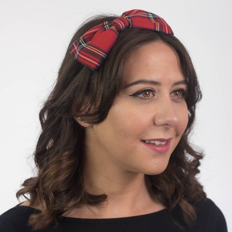May include: A red plaid headband with a bow detail. The headband features a classic tartan pattern in red, green, yellow, and blue. The accessory is worn on the head and is set against a white background.