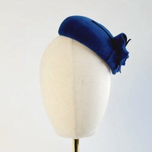 May include: A blue felt hat with a flower accent. The hat is on a white mannequin head.
