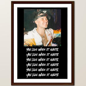 May include: Framed print featuring a smiling person wearing a headset and colorful shirt. Below the image, the phrase "YOU LIKE WHEN IT HURTS" is repeated in white text against a black background. The frame is dark brown.