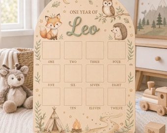 May include: An arched wooden milestone board featuring the text ONE YEAR OF Leo, with illustrations of a fox, owl, hedgehog, teepee, and campfire. The board includes twelve empty square frames labeled with the numbers one through twelve.