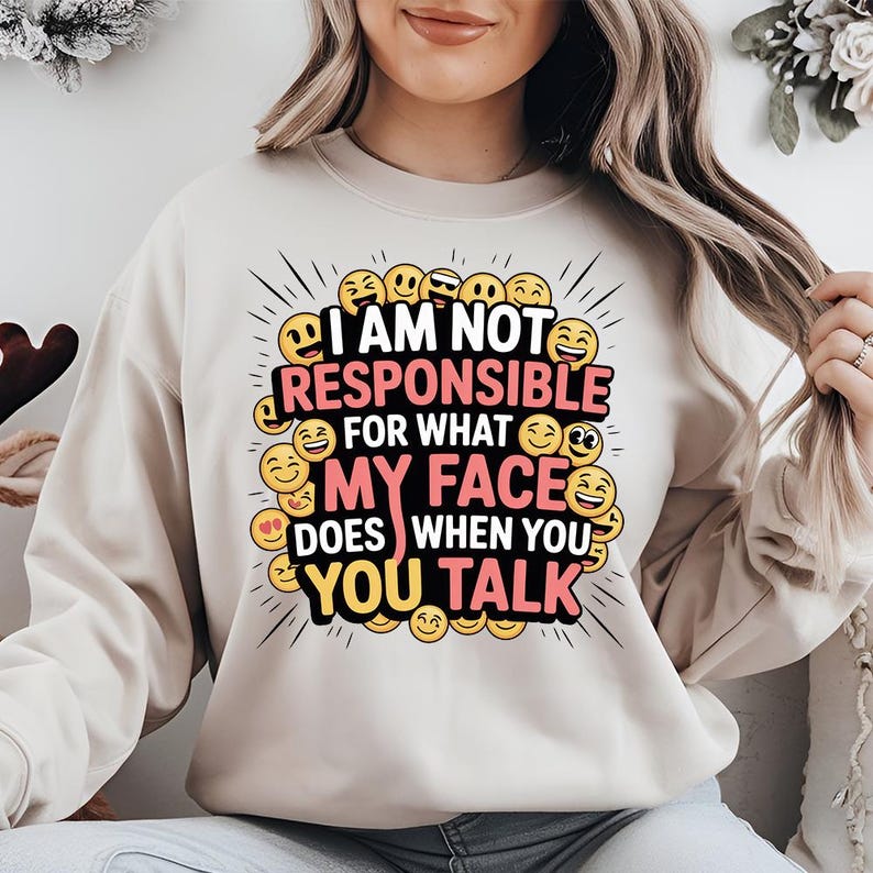 May include: Beige sweatshirt with the text "I AM NOT RESPONSIBLE FOR WHAT MY FACE DOES WHEN YOU TALK" in black, red, and pink. The design includes various emoji faces in yellow and black, with radiating lines.