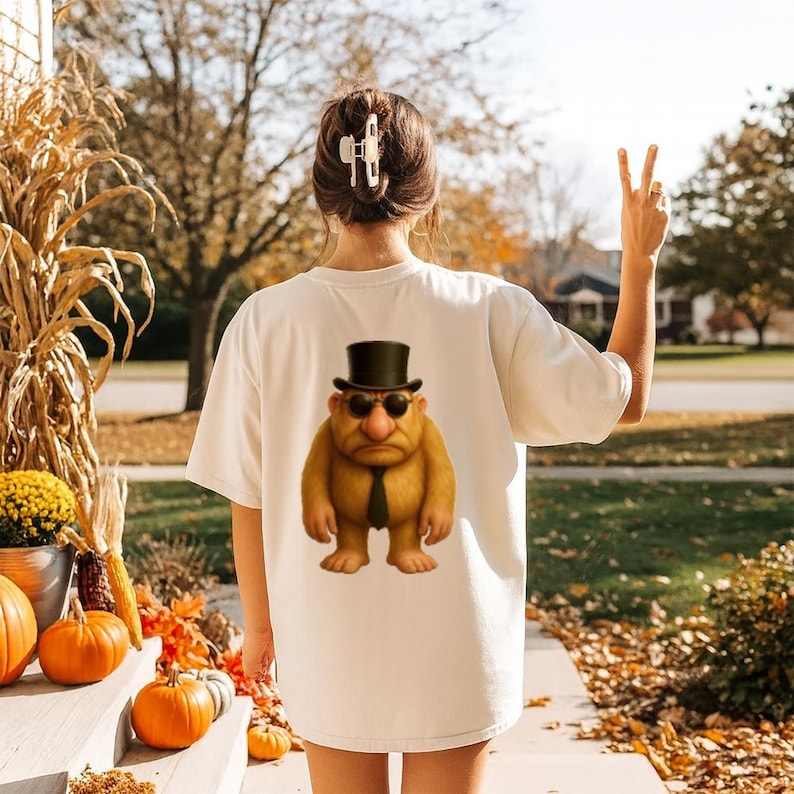 May include: A cream-coloured t-shirt with a cartoon troll wearing a top hat, sunglasses, and a black tie. The image is on the back of the shirt. The person is outside on an autumn day with pumpkins and corn stalks.