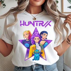 May include: White t-shirt featuring a graphic of three stylized women inside a purple star, with the word "HUNTR/X" above. The graphic includes a blue cat and the text "ALL NIGHT MINT".