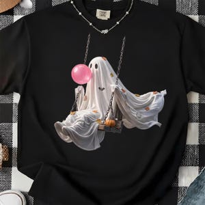May include: A black short-sleeved t-shirt featuring a graphic of a white ghost sitting on a wooden swing. The ghost is blowing a pink bubblegum bubble and is surrounded by small orange autumn leaves, a tiny bat, and a small orange pumpkin.