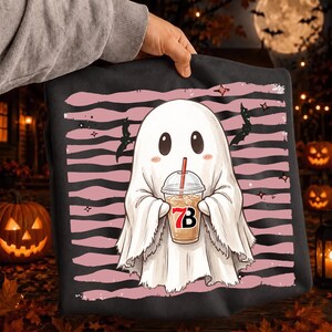 May include: A black t-shirt featuring a graphic of a white ghost with rosy cheeks holding a plastic iced coffee cup with a red straw. The cup displays the text 7B. The background of the graphic consists of horizontal pink and black stripes with bats.