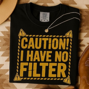 May include: Black t-shirt with the text "CAUTION! I HAVE NO FILTER" in yellow. The design includes yellow hazard symbols and a yellow border. A gold necklace is draped on the shirt.