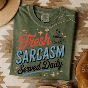 May include: Olive green t-shirt with the phrase "Fresh Sarcasm Served Daily" in red, blue, and black lettering. The shirt features a coffee cup graphic and star accents. A gold necklace rests on the shirt.
