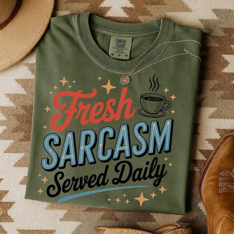 May include: Olive green t-shirt with the phrase "Fresh Sarcasm Served Daily" in red, blue, and black lettering. The shirt features a coffee cup graphic and star accents. A gold necklace rests on the shirt.